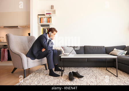 Businessman sitting on armchair using laptop and making smartphone call Stock Photo