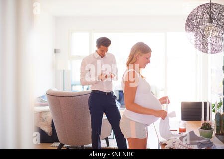 Man looking at smartphone and pregnant girlfriend folding laundry in living room Stock Photo