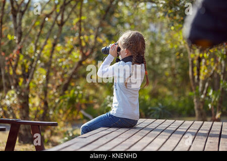 Girl in rural setting, sitting on decking, looking through binoculars Stock Photo