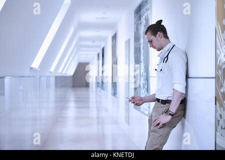 Doctor in hospital corridor leaning against wall using smartphone Stock Photo