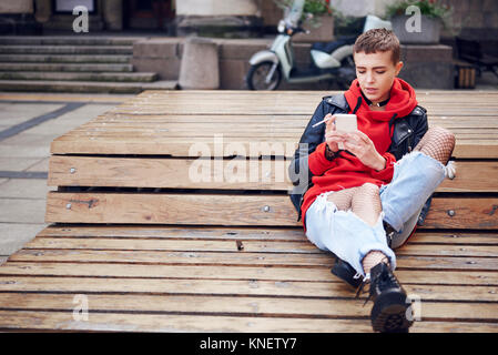 Cool young woman with short hair looking at smartphone and smoking cigarette on city bench Stock Photo