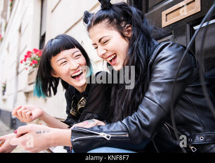 Two young stylish female friends sitting doorstep laughing Stock Photo