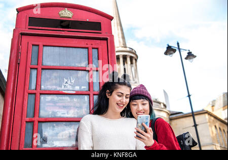 Two young stylish women looking at smartphone by red phone box, London, UK Stock Photo