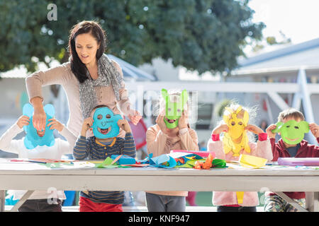Mid adult woman helping children with crafting activity, children wearing paper masks Stock Photo