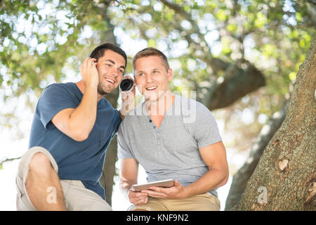 Two young male friends sitting under tree listening to headphones Stock Photo