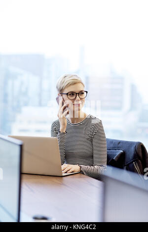 Caucasian businesswoman sat in an office using a laptop and speaking on a cell phone with copy space Stock Photo