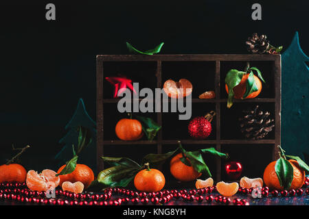Christmas tangerines in a wooden shadow box on a dark background. Vibrant ripe fruit concept with pine cones, decorations, and garlands. New Year prep Stock Photo