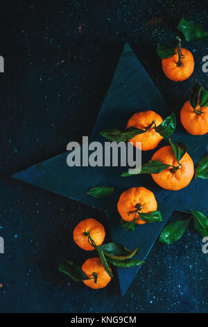 Orange tangerines with green leaves on a star-shaped cutting board. Vibrant fruits on a dark background. Rustic food photography. Stock Photo