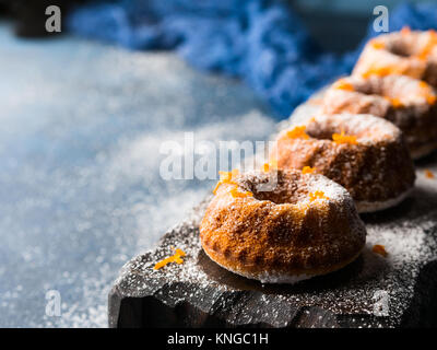 Mini bundt ring cakes with orange zest icing sugar on dark blue background and serving board. Holiday sweet food Stock Photo