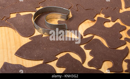 Close-up of cutting out Christmas gingerbreads on pastry board. Metallic cutter in horseshoe shape, different shapes of cookies from rolled out dough. Stock Photo