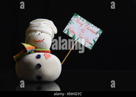 Snowman figure holding a placard board. Text “Merry Christmas & Happy New Year!” written on it, and surrounded with fir branches. Low key photograph. Stock Photo