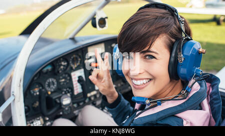 Smiling female pilot in the light aircraft cockpit, she is wearing aviator headset and making a V sign Stock Photo