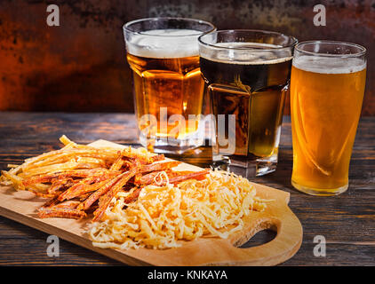 Three glasses with light, unfiltered and dark beer stand in a row near wooden cutting board with snacks on dark desk. Food and beverages concept Stock Photo