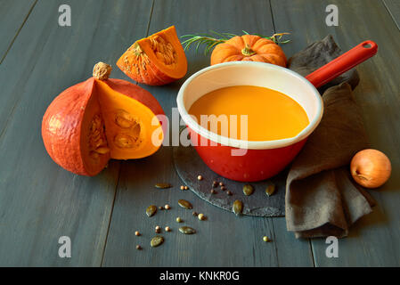 Freshly cooked pumpkin cream soup in red enamel pot on dark wooden background with cut pumpkins and a towel. Stock Photo