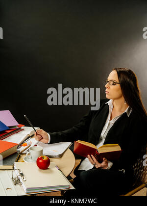 Photo of a teacher or business woman in her 30's sitting at a desk in front of a large blackboard reading and writing. Stock Photo