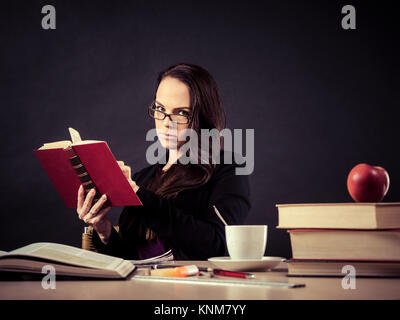 Photo of a teacher in her 30's sitting at a messy desk in front of a large blackboard reading a book. Stock Photo