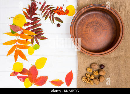 Scattered autumn leaves on white desk, empty clay plate near walnuts and chestnuts on linen cloth. Thanksgiving day and autumn concept Stock Photo