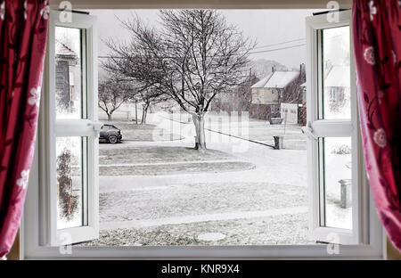 View through open window onto a beautiful winter snow street scene in rural England. Red curtains hang in front of the modern double glazed window Stock Photo