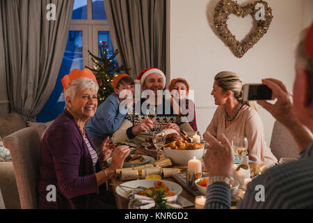 Family are having Christmas dinner together. The grandfather is using a smartphone to take a photo of his family. Stock Photo