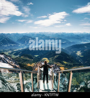 Young man with backpack stands on observation deck of skywalk rope bridge Dachstein Mountains and enjoys the landscape in Austria Stock Photo