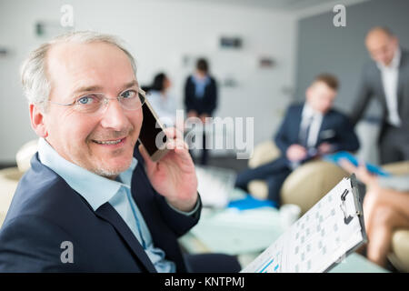 Portrait of smiling senior businessman talking on smartphone with colleagues in background at office Stock Photo