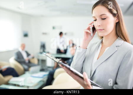Businesswoman reading document on clipboard while using smartphone in meeting room at office Stock Photo
