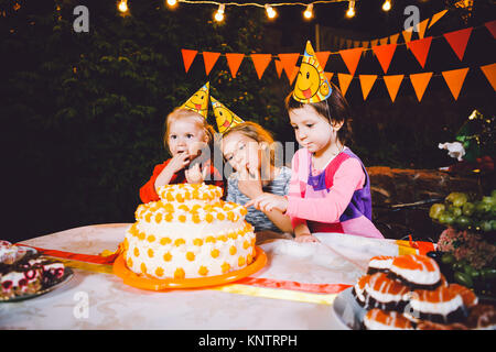 Children's birthday party. Three cheerful children girls at the table eating cake with their hands and smearing their face. Fun and festive mood in the decorated courtyard decor with bright bulbs Stock Photo