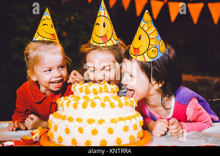 Children's birthday party. Three cheerful children girls at the table eating cake with their hands and smearing their face. Fun and festive mood in the decorated courtyard decor with bright bulbs Stock Photo