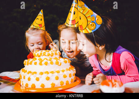 Children's birthday party. Three cheerful children girls at the table eating cake with their hands and smearing their face. Fun and festive mood in the decorated courtyard decor with bright bulbs Stock Photo