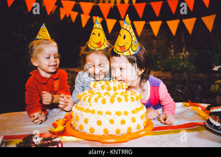 Children's birthday party. Three cheerful children girls at the table eating cake with their hands and smearing their face. Fun and festive mood in the decorated courtyard decor with bright bulbs Stock Photo