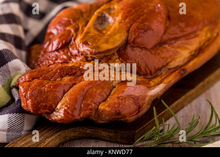 Horizontal photo with big piece of smoked pork meat with bone placed on wooden cutting board. Blue checkered towel and green rosemary herb are around  Stock Photo