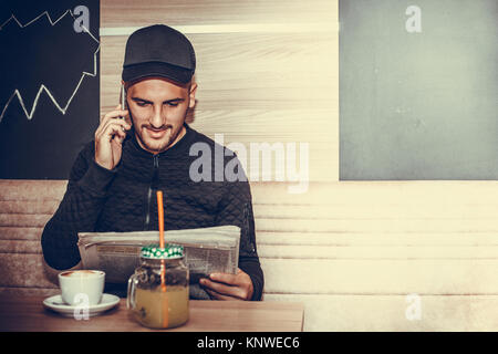 Young man with cap reading the newspaper, talking on a mobile phone and drinking coffee in a coffee shop. Student / sportsman reading the latest sport Stock Photo