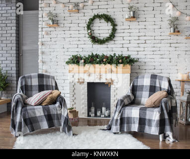 Christmas festive interior in grey and white colors. Round green wreath hanging on white brick wall. Two armchairs covered with cozy blankets standing Stock Photo