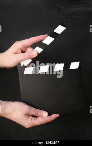 A blank clapperboard held open as if about to snap shut between the hands of a male operative with black chalkboard background. Stock Photo