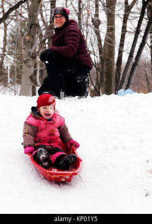 A little girl (3 yr old) sledging in Quebec, while mum looks on Stock Photo