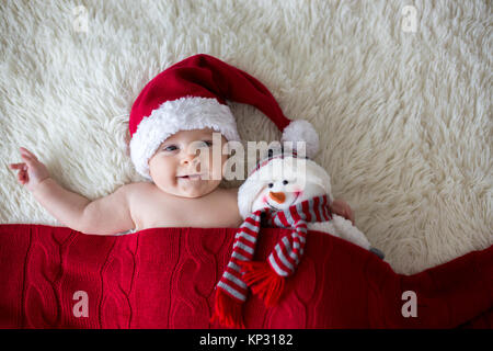 Christmas portrait of cute little newborn baby boy, wearing santa hat and hugging little cute snowman toy, studio shot, winter time Stock Photo