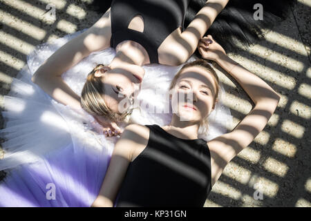 Ballet dancers lying on the tutus on the concrete floor outdoors. They wears a black dance wear. Girls looking into the camera. Sunlight creates strip Stock Photo