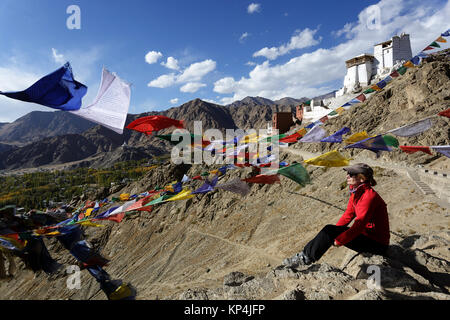 Woman in red jacket sitting on a rock under prayer flags with Tsemo Gompa in the background, Leh, Ladakh, Jammu and Kashmir, India. Stock Photo