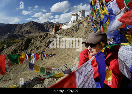 A close-up of a woman in red jacket sitting on a rock under prayer flags with Tsemo Gompa in the background, Leh, Ladakh, Jammu and Kashmir, India. Stock Photo