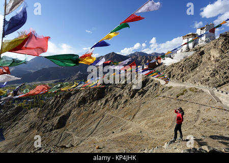 Woman in red jacket standing on a rock under prayer flags with Tsemo Gompa in the background, Leh, Ladakh, Jammu and Kashmir, India. Stock Photo