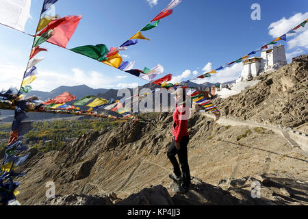 Woman in red jacket standing on a rock under prayer flags with Tsemo Gompa in the background, Leh, Ladakh, Jammu and Kashmir, India. Stock Photo
