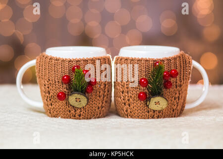 Close up of two white mugs in cute brown knitted sweaters with christmas decorations at blurry lights of orange garland background. Stock Photo