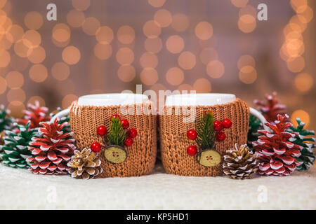 Closeup of two white cups in brown knitted sweaters with christmas decorations at lights of orange garland background Stock Photo