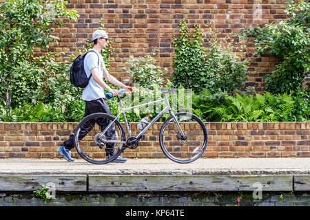 Young man in baseball cap and trainers pushing a bike along the towpath of Regent's Canal at King's Cross, London, UK, on a summer afternoon Stock Photo
