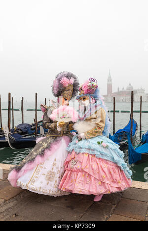 Venice Carnival 2017, Veneto, Italy. Two women in pink  costumes and masks posing together in front of gondolas on a misty high key day Stock Photo