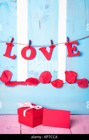 Cute red gift box with a blank message card near it and the word love written in red paper letters tied to a string, on a blue wooden fence. Stock Photo