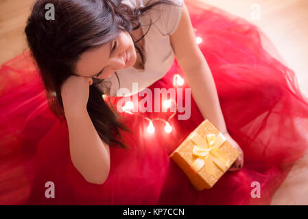 Beautiful young brunette woman, dressed in a red tutu, sitting on the floor, surrounded by Christmas lights and holding a gift box with a tied bow. Stock Photo