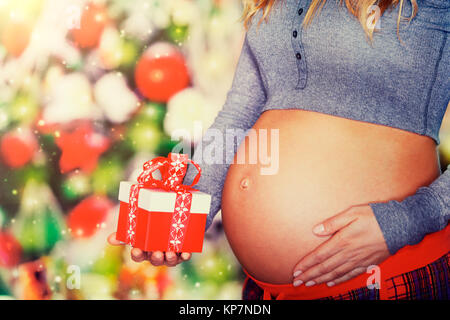 Closeup photo of a tummy of pregnant woman with gift box in hand on festive decorated fir tree background, body part, best present on Christmas for yo Stock Photo