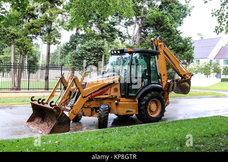 Yellow cabbed tractor with backhoe and muddy front loader on an upscale neighborhood street on a rainy day Stock Photo