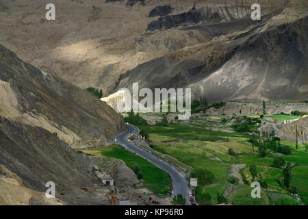 A wide mountain road, gray asphalt passes through a green valley among the rocks Moonland, the slopes of the mountains are lit by the rays of the sett Stock Photo